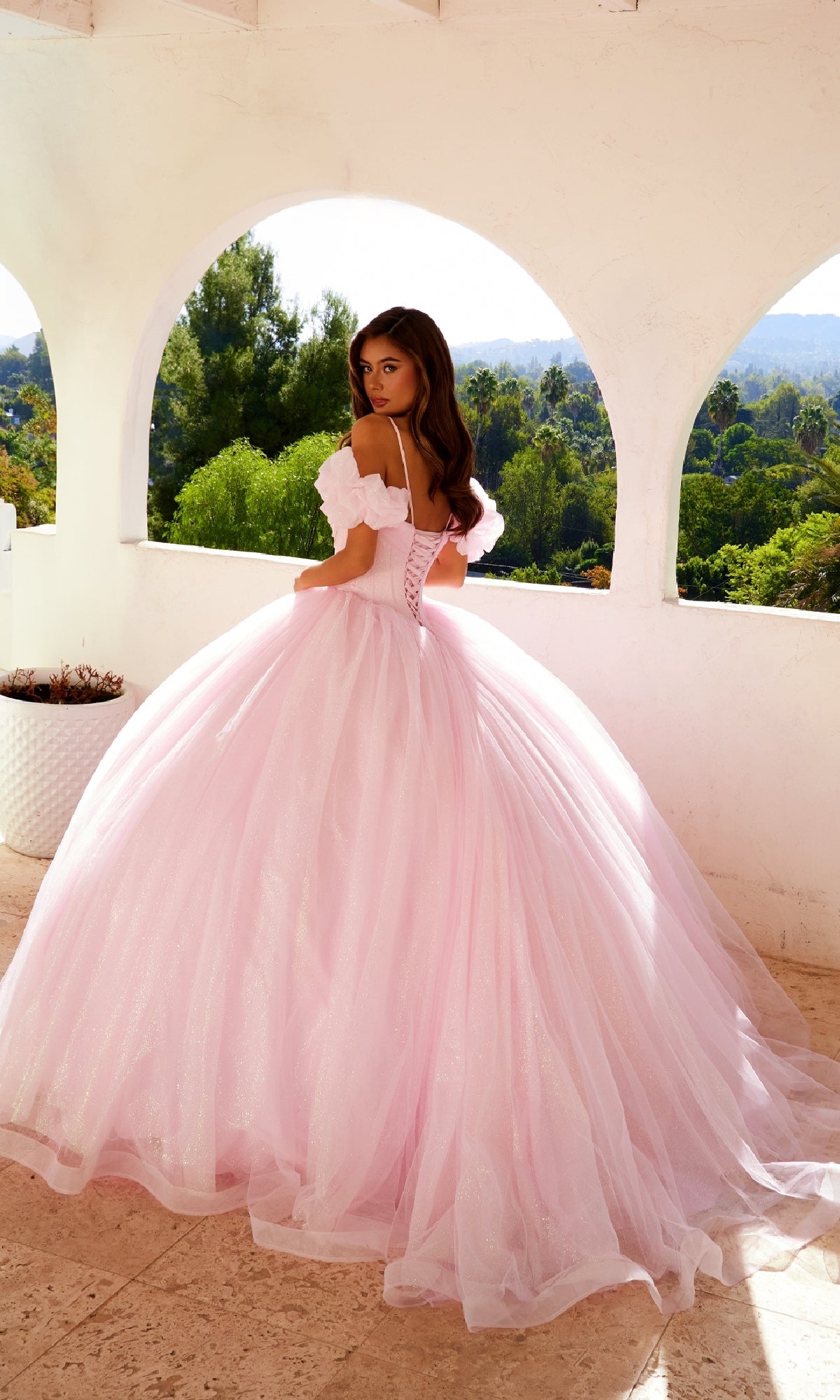 A woman wearing the Quinceanera Dress JT1487H By Juliet, a glitter tulle ball gown with puff off-the-shoulder sleeves, stands on a balcony with white arches overlooking lush greenery and distant hills.